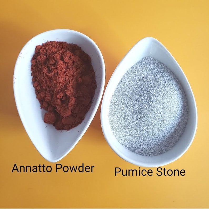 May include: Two white bowls on a yellow background. The bowl on the left contains a red powder labeled "Annatto Powder". The bowl on the right contains a light gray powder labeled "Pumice Stone".