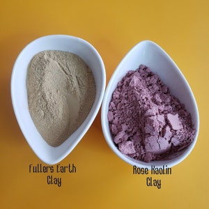 May include: Two white bowls with different types of clay. The bowl on the left contains a light brown clay labeled "Fullers Earth Clay". The bowl on the right contains a pink clay labeled "Rose Kaolin Clay".