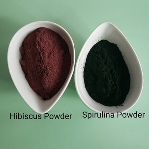 May include: Two white bowls, one containing a red powder labeled "Hibiscus Powder" and the other containing a dark green powder labeled "Spirulina Powder".