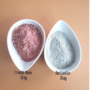 May include: Two white bowls, one containing pink French Rose Clay and the other containing gray Bentonite Clay.