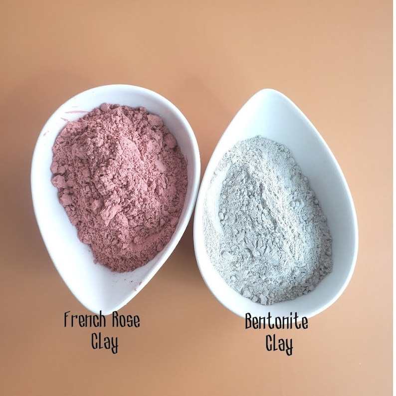 May include: Two white bowls, one containing pink French Rose Clay and the other containing gray Bentonite Clay.
