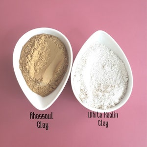 May include: Two white bowls filled with different types of clay. The bowl on the left contains a light brown clay labeled "Rhassoul Clay". The bowl on the right contains a white clay labeled "White Kaolin Clay".