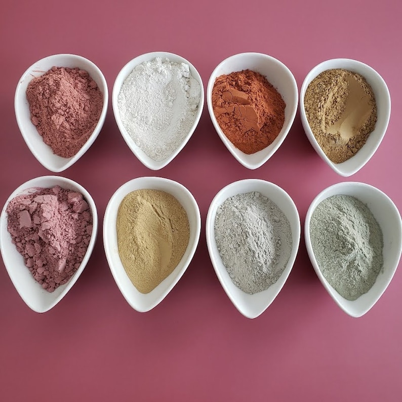 May include: Eight small white bowls filled with different colored clay powders. The powders are pink, white, red, brown, light pink, beige, gray, and green.