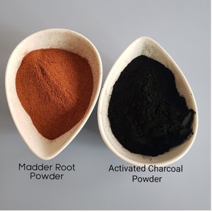 May include: Two small white bowls, one containing a pile of red-brown powder labeled "Madder Root Powder" and the other containing a pile of black powder labeled "Activated Charcoal Powder".