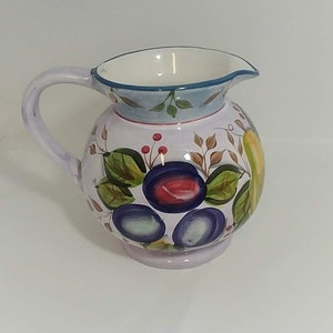 May include: A white ceramic pitcher with a light blue rim and a floral design featuring purple, red, green, and yellow accents. The pitcher has a round body and a handle.