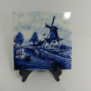 May include: Blue and white ceramic tile depicting a Dutch windmill scene with a canal, bridge, and houses. The tile is on a black easel.