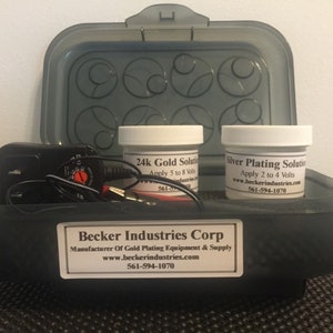 May include: A black plastic container with two white jars labeled "24k Gold Solution" and "Silver Plating Solution". The jars are next to a black power adapter with wires. The container has a label that reads "Becker Industries Corp. Manufacturer of Gold Plating Equipment & Supply www.beckerindustries.com 561-594-1070".