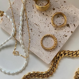 May include: A set of gold jewelry featuring a chunky chain necklace, a pearl necklace, and four gold rings. The rings are different styles, including a thick band, a twisted band, and a simple band.