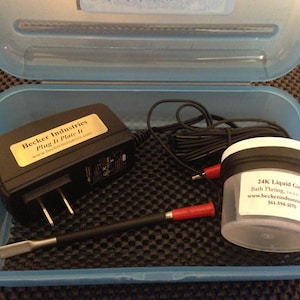 May include: A blue plastic container with a black power adapter, a small clear jar with a black lid, and a black metal tool. The jar is labelled "24K Liquid Gold Bath Plating, Use 1 oz per 50 gal" and includes the website address "www.beckerindustries.com" and phone number "561-594-8070".