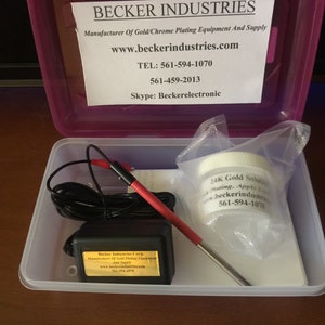 May include: A white plastic container with a clear lid. Inside the container is a black and red electrical cord, a small black box with a gold label, and a white bag with the text "24K Gold Solution Plating, Apples, beckerindustries.com, 561-594-1079".