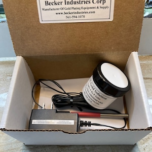 May include: A white box with a black power adapter, a black and white jar labeled "24K Gold Solution", and a red and silver probe. The box has a label that reads "Becker Industries Corp. Manufacturer of Gold Plating Equipment & Supply www.beckerindustries.com 561-594-1070".