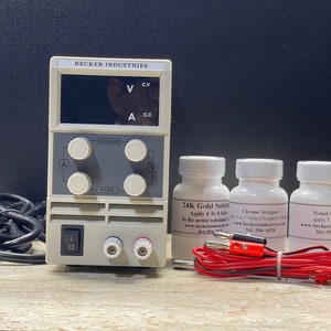 May include: A white power supply unit with a digital display, two knobs, and a power switch. Three white bottles labelled "24K Gold Solution", "Chrome Stripper", and "Nickel Activator" are in the background. Red and black electrical wires are in the foreground.