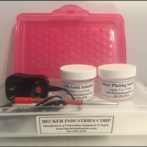 Puede incluir: Un kit de plateado en oro en un recipiente de plástico transparente con tapa rosa. El kit incluye dos frascos blancos etiquetados como "24k Gold Solution" y "Silver Plating Solution", un adaptador de corriente negro, un cable rojo y negro y una herramienta de metal plateado. El recipiente tiene una etiqueta en la parte inferior que dice "BECKER INDUSTRIES CORP. Fabricante de equipos y suministros de plateado en oro www.beckerindustries.com 561-594-1070"
