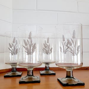 May include: Set of four clear glass stemmed drinking glasses with a wheat design. The glasses have square bases and are a dark grey colour.