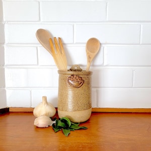 May include: A speckled tan ceramic utensil holder with a decorative design, holding a wooden fork and spoon. A whole garlic bulb, a garlic clove, and green leaves are on the wooden surface. The background is a white brick wall.