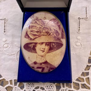 Peut inclure: Une boîte à bijoux ovale vintage avec un intérieur en velours bleu. Le couvercle présente une image peinte d'une femme portant un grand chapeau à larges bords avec un nœud violet.