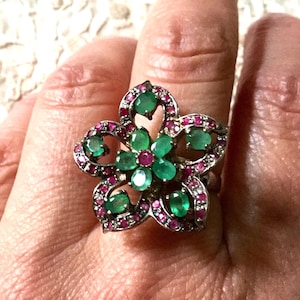 Peut inclure: Une bague en forme de fleur ornée de pierres précieuses vertes et rouges. La bague présente un anneau de couleur argentée et est ornée de pierres de couleur émeraude au centre et autour des pétales, avec de plus petites pierres rouges bordant les pétales.