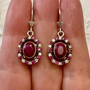 Op de afbeelding: Een paar sierlijke oorbellen met ovale, dieprode edelstenen in het midden, omringd door kleinere roze en witte stenen. De oorbellen hebben zilverkleurige haken en decoratieve bovenkanten.