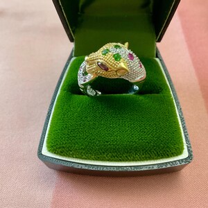 Peut inclure: Bague en or et argent en forme de tête de jaguar, ornée de pierres précieuses vertes, rouges et blanches. La bague est présentée dans un écrin noir avec un intérieur en velours vert. Le texte est visible en haut.