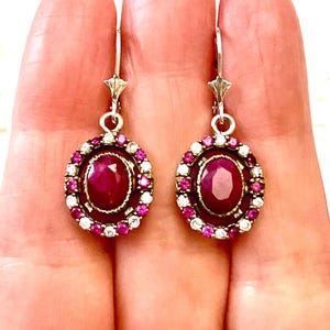 Peut inclure: Une paire de boucles d'oreilles ovales avec une pierre centrale rouge foncé, entourée de petites pierres roses et blanches. Les boucles d'oreilles ont une fermeture à crochet argentée. Bijoux classiques.