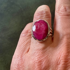 Peut inclure: Une bague en argent avec une grande pierre précieuse de rubis facetté de forme ovale. La bague a une bande texturée.