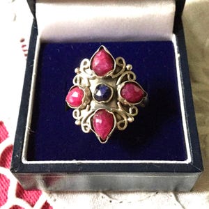Peut inclure: Bague en argent avec un motif floral, sertie de cinq pierres rouges et d'une pierre bleue au centre. La bague est présentée dans un écrin en velours bleu.