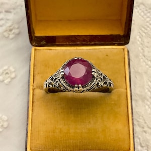 Op de afbeelding: Vintage zilveren ring met een grote, ronde, gefacetteerde robijn. De ring heeft een sierlijk filigraanontwerp rond de edelsteen. De ring wordt getoond in een vintage sieradendoosje met de tekst "AU ROY YVETOT ROUEN".