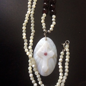 Peut inclure: Un collier avec un pendentif en forme de coquille blanche, orné d'une croix sculptée et d'une pierre précieuse rouge au centre. Le collier est composé de perles blanches et possède un fermoir argenté.