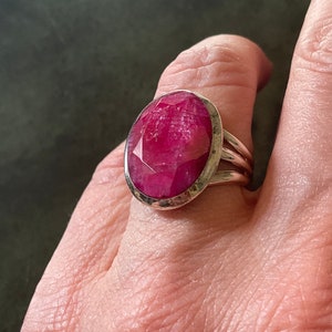 Peut inclure: Une bague en argent avec une grande pierre précieuse de rubis de forme ovale facetée. La bague a une bande simple et polie.