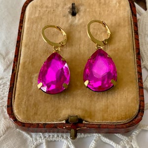 May include: A pair of magenta teardrop earrings with gold-coloured leverback closures. The earrings are displayed in a vintage, red jewellery box with a tan interior. The earrings have a faceted, jewel-like appearance.