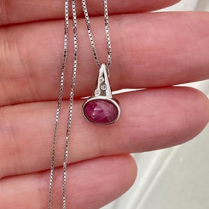 Peut inclure: Un collier pendentif en argent avec une pierre précieuse rubis. Le pendentif a un sommet triangulaire avec trois petites pierres claires et un rubis ovale. La chaîne est une chaîne délicate.