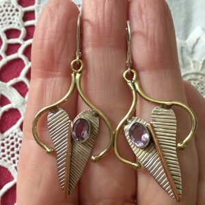 May include: A pair of silver and gold-toned earrings with a leaf design. Each earring features a central oval amethyst stone and a textured silver leaf shape with a copper accent. The earrings have a hook closure.