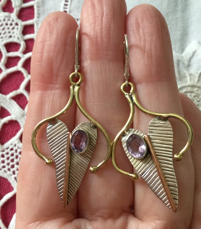 May include: A pair of silver and gold-toned earrings with a leaf design. Each earring features a central oval amethyst stone and a textured silver leaf shape with a copper accent. The earrings have a hook closure.