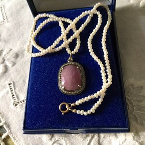 Peut inclure: Un pendentif en pierre rose avec un halo de diamants suspendu à un collier de perles blanches. Le collier est présenté dans un écrin en velours bleu.