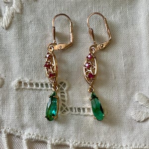 Op de afbeelding: Een paar goudkleurige oorbellen met een klapsluiting. Elke oorbel heeft een ovaal ontwerp met rode edelstenen en een druppelvormige, smaragdgroene edelsteenhanger. De oorbellen worden gepresenteerd op een wit, getextureerd oppervlak.