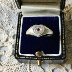 Peut inclure: Une bague en argent avec un motif en forme de cœur représentant une rose en relief avec une pierre précieuse rouge au centre. La bague est dans un écrin à bague en velours noir.