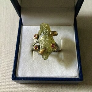 Peut inclure: Une bague ornée d'une grande pierre brute translucide vert clair, montée sur un anneau en métal sombre et texturé. Le design comprend trois petites pierres rouges rondes serties dans des griffes dorées retenant la pierre centrale.