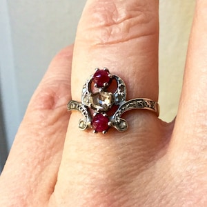 Peut inclure: Une bague en argent avec un design délicat orné de trois pierres précieuses. Deux des pierres sont rouges et une est une pierre claire et facetée.