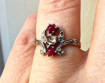 May include: A silver ring with a delicate design featuring three gemstones. Two of the stones are red and one is a clear, faceted stone.