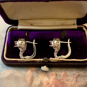 Peut inclure: Paire de boucles d'oreilles argentées avec une petite pierre rouge ronde au centre, entourée de petites pierres transparentes. Les boucles d'oreilles sont présentées dans un écrin à bijoux vintage en velours violet.