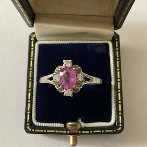 Peut inclure: Une bague en argent avec une pierre ovale rose entourée de diamants plus petits. La bague est dans un écrin à bague en velours bleu.