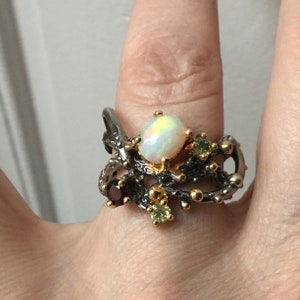 Peut inclure: Une bague en argent avec une opale blanche, entourée de petites pierres précieuses vertes et rouges, sertie dans un motif de branche torsadée.