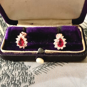 Peut inclure: Boucles d'oreilles en forme de goutte d'eau avec une pierre rouge centrale entourée de petites pierres blanches. Présentées dans un écrin de bijoux en velours violet ouvert.