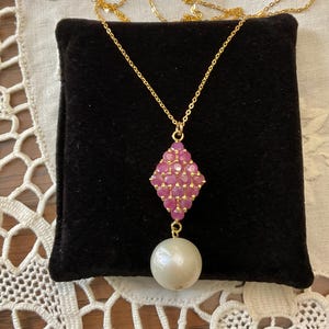 Peut inclure: Un collier doré avec un pendentif. Le pendentif présente un motif en losange de pierres précieuses roses et une grosse perle blanche ronde. Le collier est présenté sur une surface en velours noir.