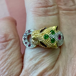 Peut inclure: Une bague dorée en forme de tête de jaguar. La bague est ornée de pierres précieuses vertes et rouges, avec une finition dorée texturée. Les yeux du jaguar sont verts et la bague a un anneau argenté avec des pierres rouges.