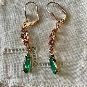 May include: A pair of gold-coloured earrings with a lever back closure. Each earring features a teardrop-shaped green gemstone and a cluster of small red gemstones. The earrings are displayed on a white, textured surface.