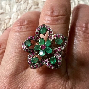 May include: A silver ring with a flower design featuring green emeralds and pink rubies.