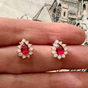 Peut inclure: Une paire de boucles d'oreilles en forme de goutte d'eau avec une pierre précieuse rouge entourée d'un halo de petites pierres blanches.