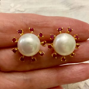 Peut inclure: Paire de boucles d'oreilles à clous en perles. Chaque boucle d'oreille présente une grosse perle blanche ronde au centre, entourée de petites pierres précieuses rouges serties dans du métal doré. Fond neutre.