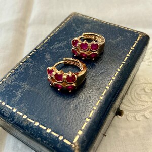 Peut inclure: Une paire de créoles dorées ornées de deux rangées de petites pierres rondes d'un rouge éclatant serties dans le métal. Les boucles d'oreilles sont de forme circulaire avec un mécanisme de fermeture à charnière, présentant une finition dorée.